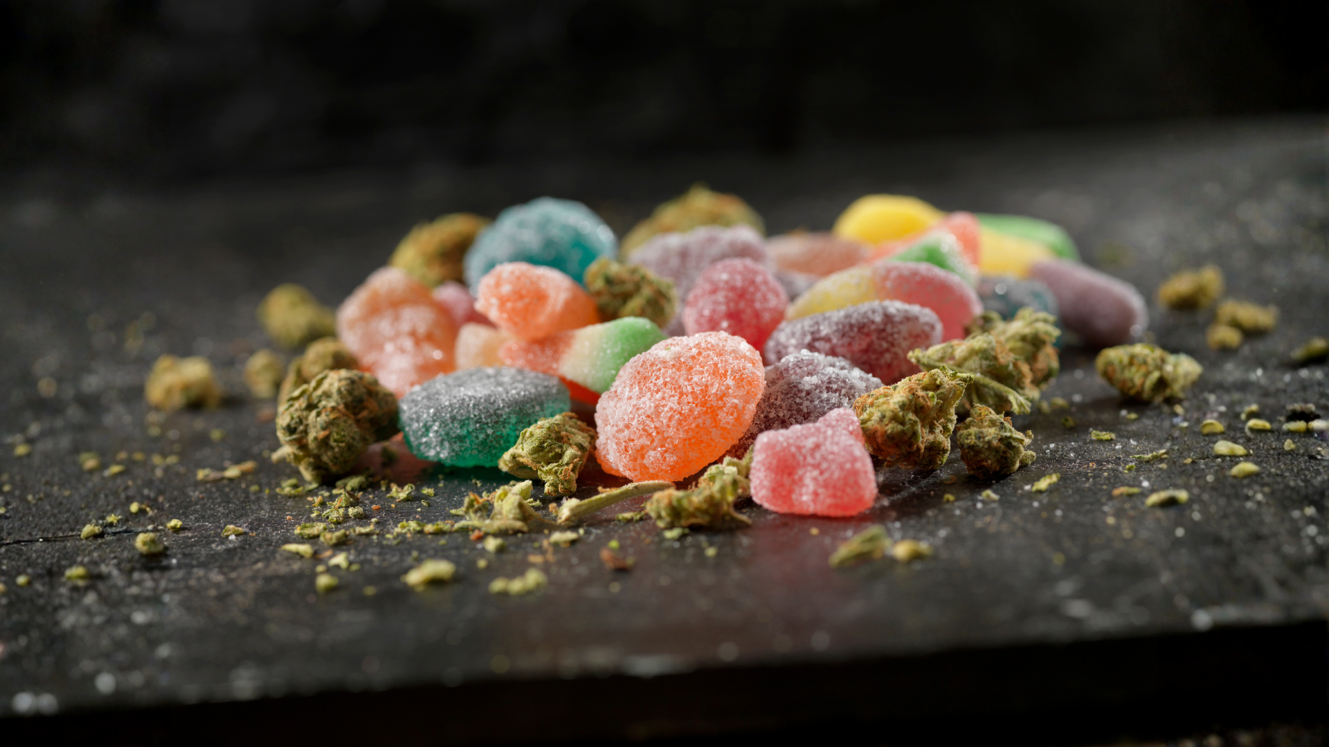 Colorful cannabis-infused gummies and marijuana buds on dark surface for edible display.