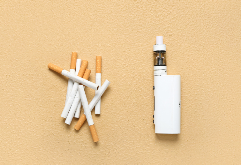 Pile of cigarettes next to white vape device symbolizing switch from smoking to vaping.